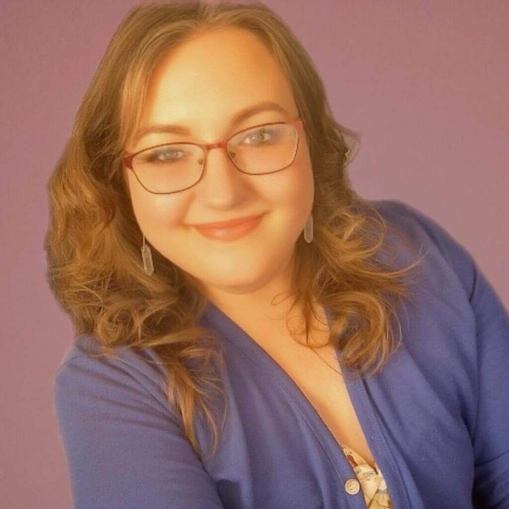 Жінка з хвилястим світло-каштановим волоссям, в окулярах і з впевненою посмішкою позує на фіолетовому тлі, одягнена в синій топ.