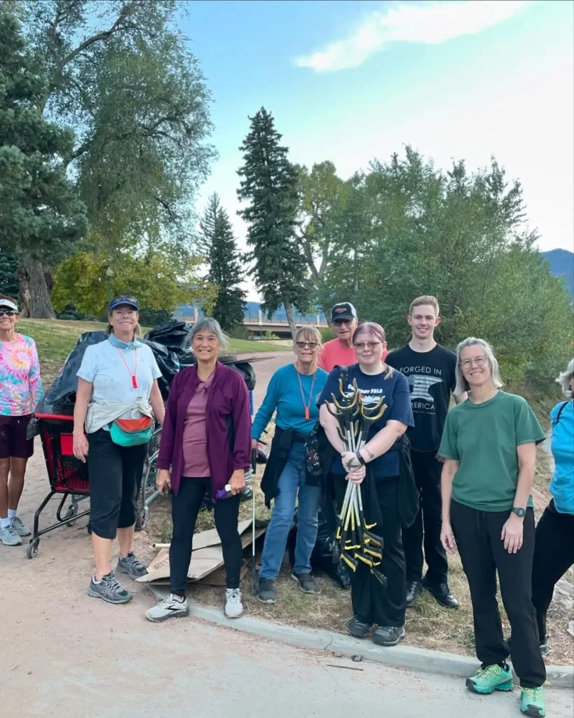 Група з десяти людей посміхається на відкритому повітрі, тримаючи садові інструменти, поруч із зібраними мішками для сміття. Вони оточені високими деревами і чистим небом, що символізує командну роботу і громадську службу.