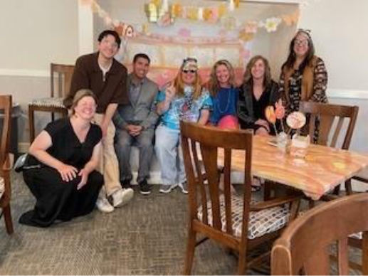 Група з семи осіб, які посміхаються і сидять у прикрашеній кімнаті з квітковими та пастельними гірляндами. Атмосфера святкова і радісна.