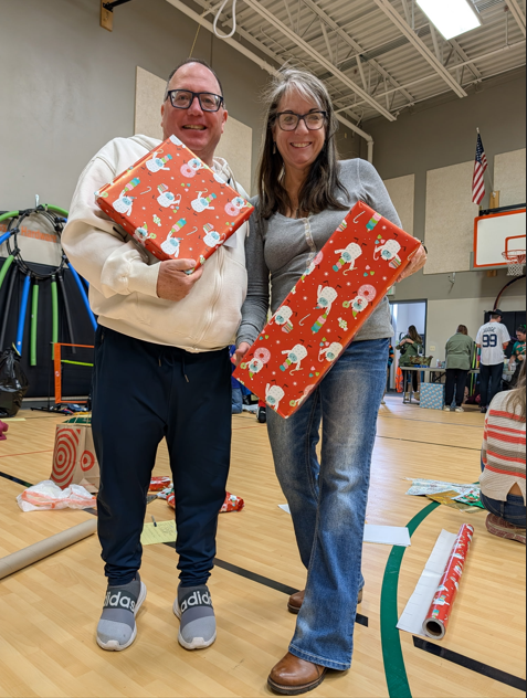 Двоє людей стоять у спортзалі, тримаючи в руках подарунки, загорнуті в червоний святковий папір. Навколо спортзалу видно пакувальні матеріали та інших волонтерів.